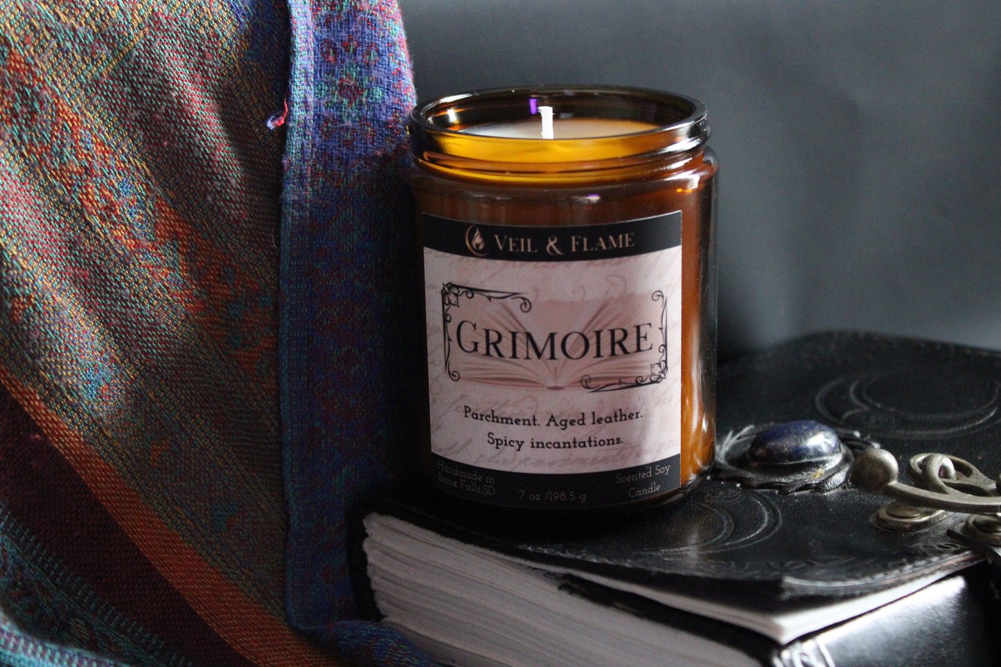 Candle in a jar labeled 'Grimoire' on a dark surface with a plaid cloth and notebook.