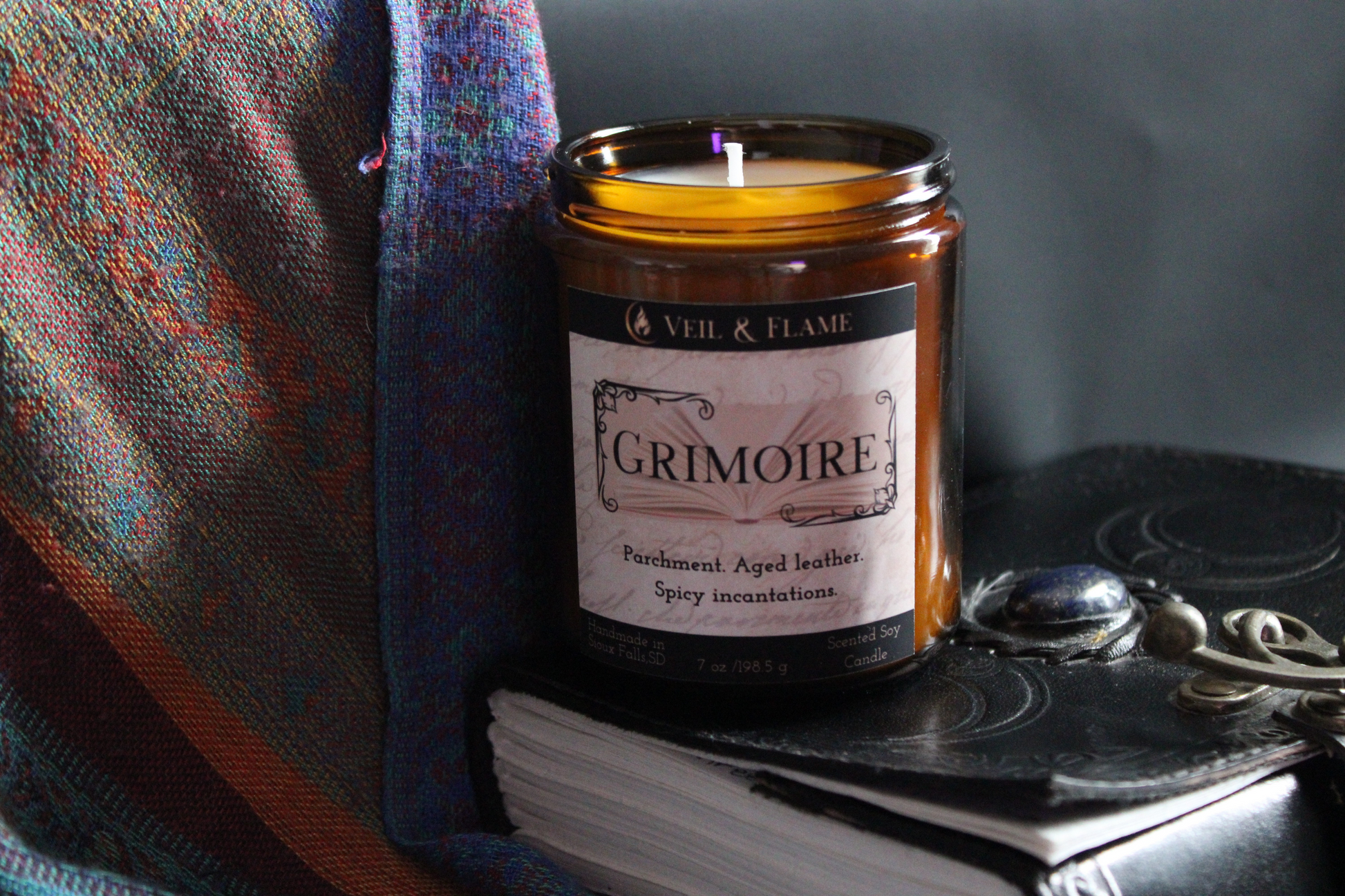 Candle in a jar labeled 'Grimoire' on a dark surface with a plaid cloth and notebook.