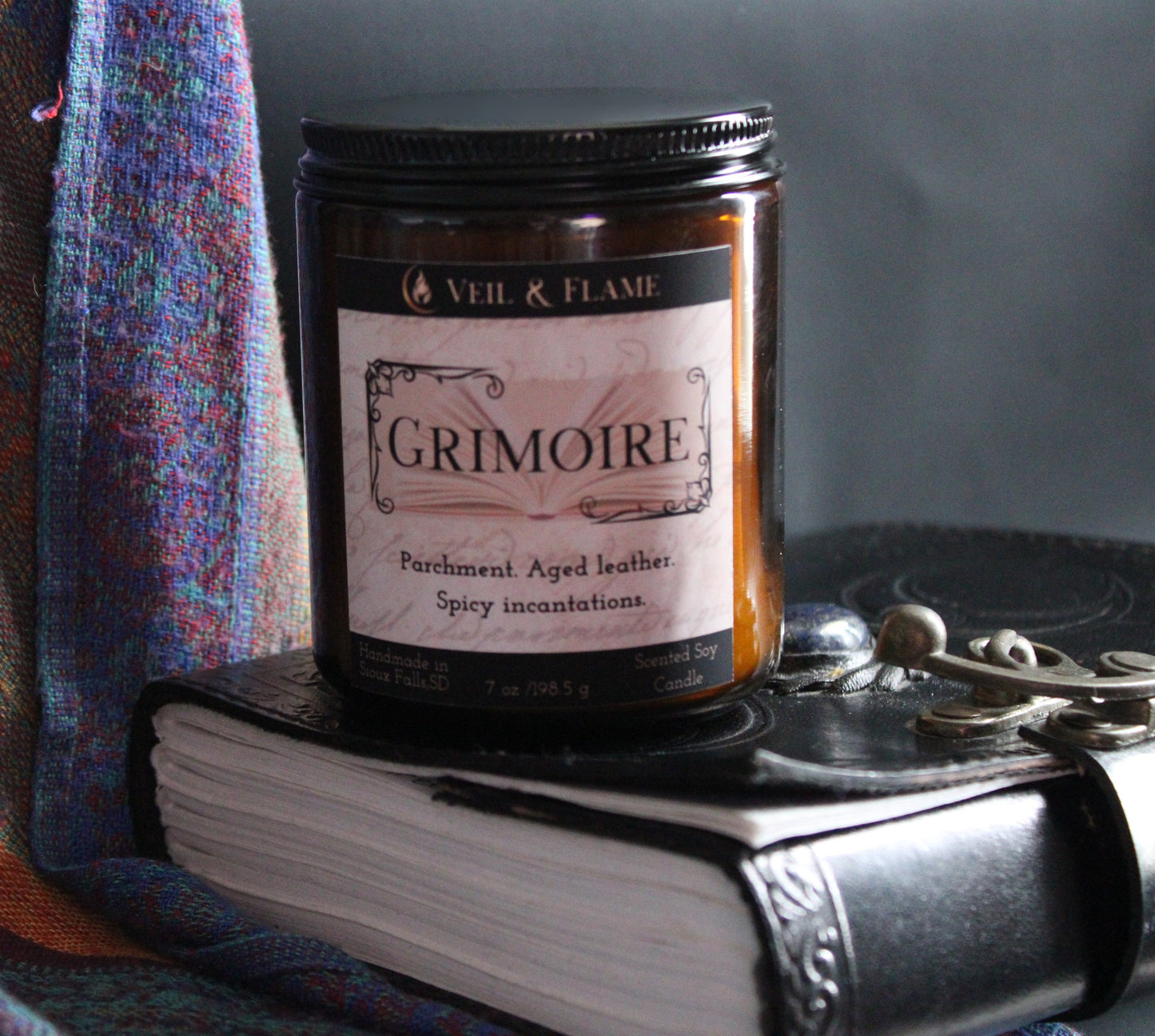 Candle labeled 'Grimoire' by Veil & Flame on top of a book with a textured fabric background.