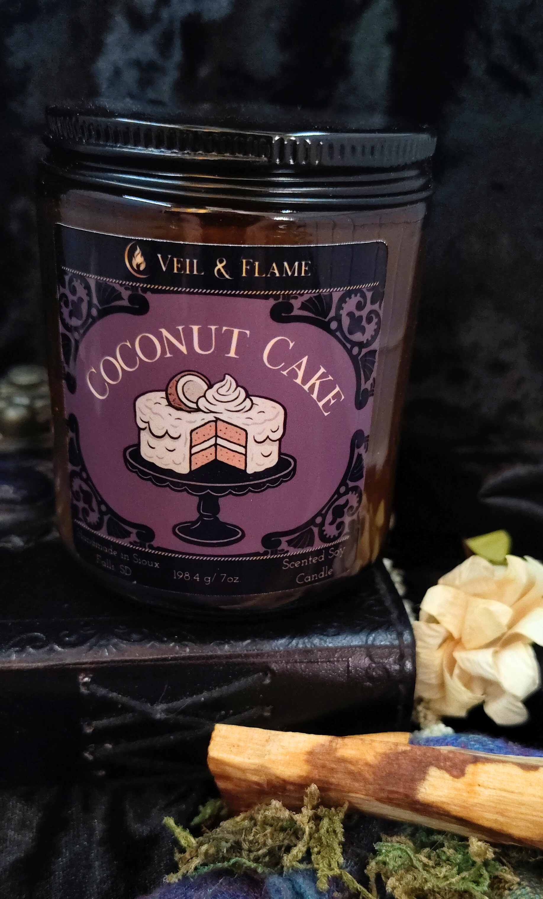 Jar of 'Coconut Cake' candle by Veil & Flame on a dark background with decorative elements.